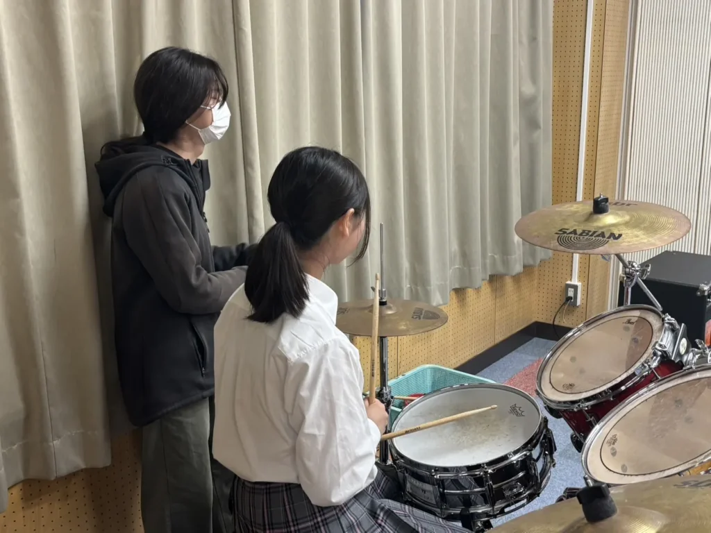 ドラムの前に座り、となりに立つ人から叩き方を教わる参加者
